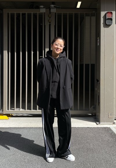 Blazer negro sobre una sudadera negra, combinado con pantalones de chándal negros con rayas blancas. Zapatillas blancas. El fondo muestra un área cerrada.