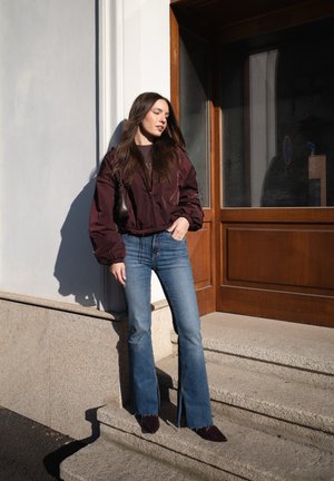 Mujer con chaqueta granate, vaqueros acampanados azules y zapatos oscuros, de pie sobre escalones de piedra junto a una puerta de madera bajo la luz del sol.