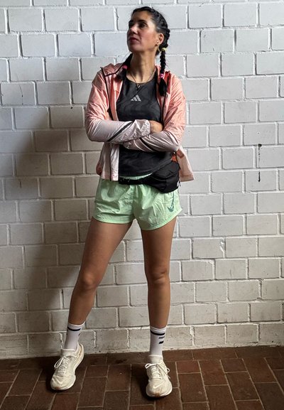 Rompevientos rosa claro, camiseta negra, pantalones cortos verde menta, zapatillas blancas y riñonera negra. Cabello trenzado. Frente a una pared de ladrillos blancos.