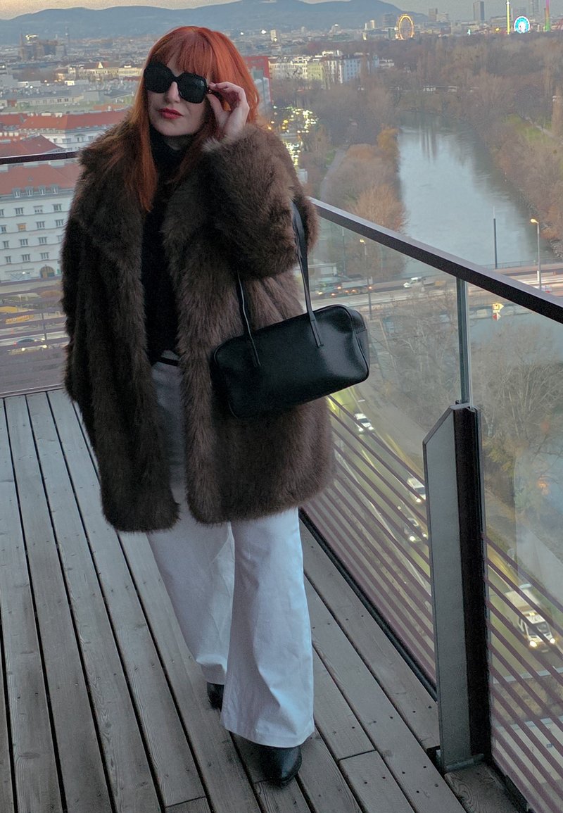 Manteau en fausse fourrure marron, haut noir et pantalon blanc à jambes larges. Sac à main noir avec bandoulière. Le mannequin porte des lunettes de soleil, debout sur une terrasse en bois.