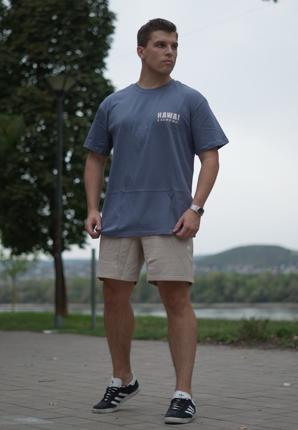 Un homme se tient dehors sur un chemin pavé, portant un t-shirt bleu Hawaii, un short beige, des baskets Adidas noires et une montre-bracelet, regardant sur le côté.