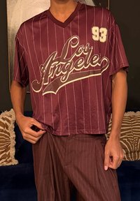 Άτομο που φοράει πουκάμισο Λος Άντζελες σε βυσσινί χρώμα με λεπτές ρίγες και αριθμό 93, καθώς και παντελόνι με τις ίδιες ρίγες, σε εσωτερικό χώρο με διακοσμητικά μαξιλάρια με σχέδια στο φόντο.