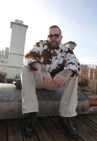 Homme barbu tatoué portant des lunettes de soleil, une veste en fourrure, un pantalon beige et des chaussures noires, assis sur un toit avec une cheminée et un plancher en bois.