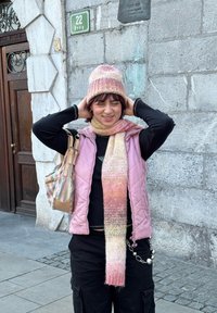 Bonnet rayé rose tricoté et crème, écharpe assortie, et une veste matelassée rose pâle portée sur un haut noir, en contraste avec un pantalon cargo noir.