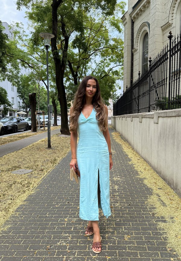 Una mujer con un vestido azul claro está de pie en una acera urbana flanqueada por árboles, junto a un edificio de piedra ornamentado y una verja de hierro.