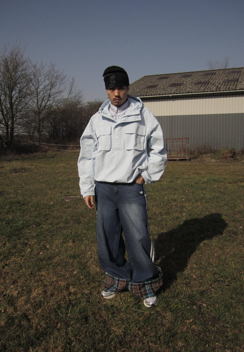 Man in oversized light jacket, beanie, and baggy jeans standing on grass near a barn under clear sky.
