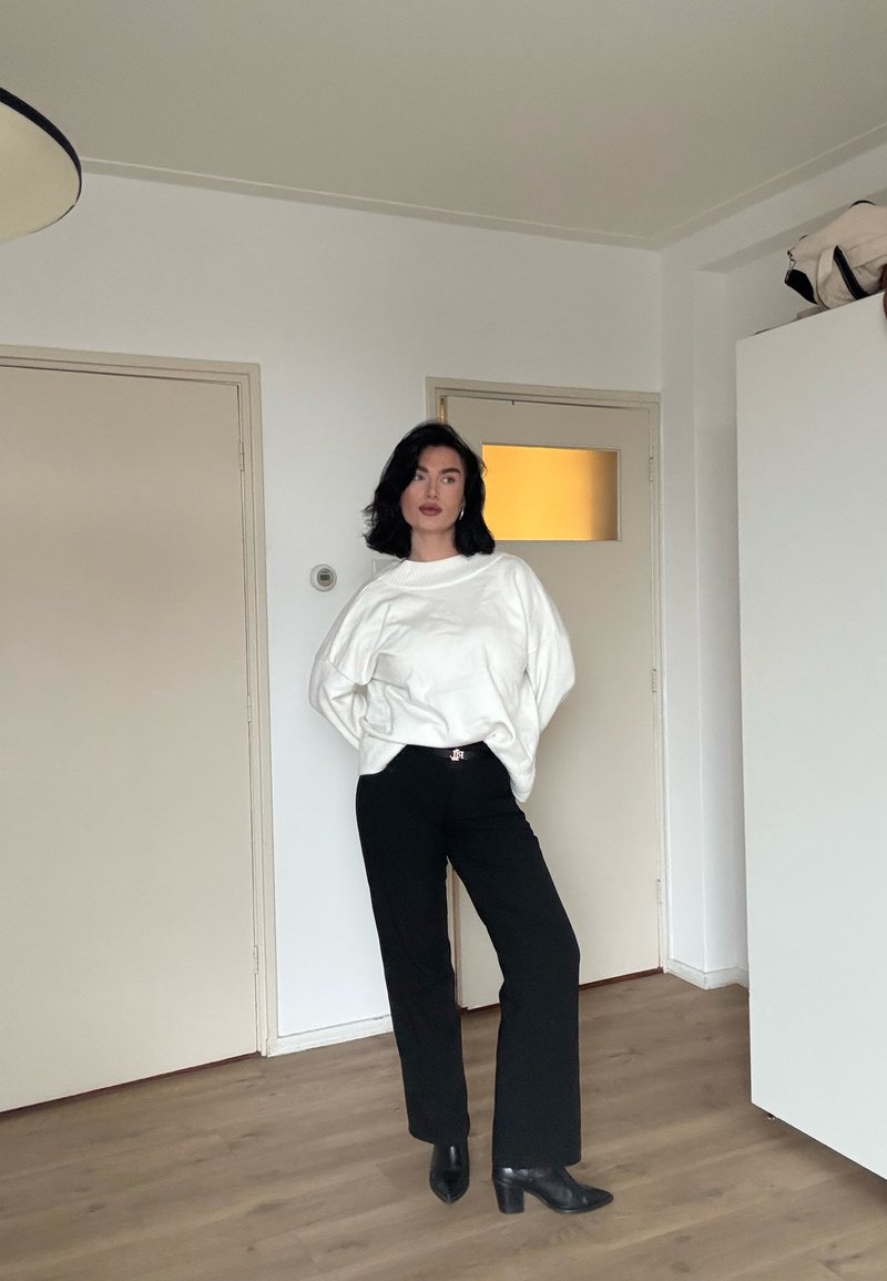Femme aux cheveux noirs portant un pull blanc surdimensionné, un pantalon noir et des bottes à talons noires, debout dans une pièce minimaliste avec un sol en bois.