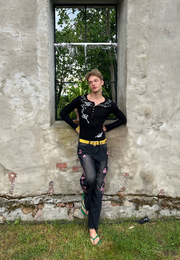 Personne appuyée contre un mur en pierre sous une fenêtre, portant un haut noir brodé, un pantalon à motifs floraux, une ceinture jaune et des sandales vertes.