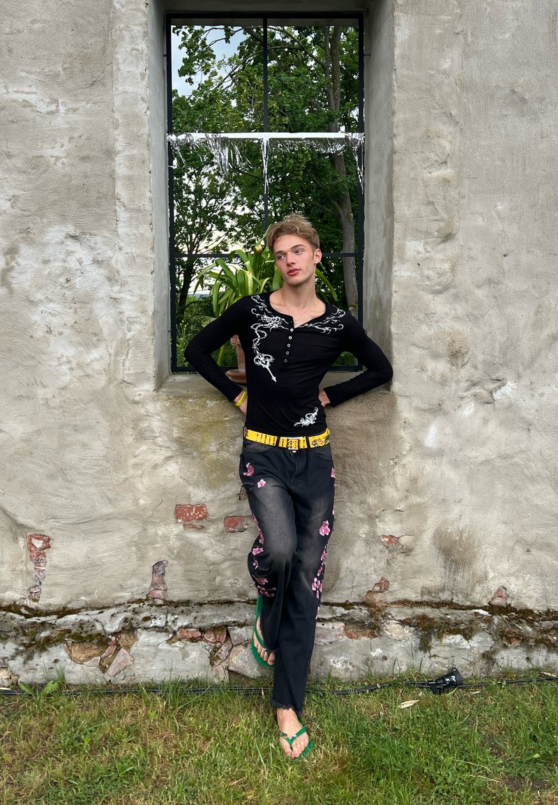 Personne appuyée contre un mur en pierre sous une fenêtre, portant un haut noir brodé, un pantalon à motifs floraux, une ceinture jaune et des sandales vertes.