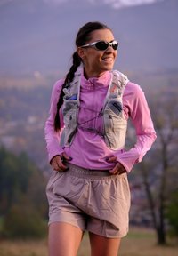 Moteris su akiniais nuo saulės, vilkinti hidraciniu liemeniu ir rožine ilgomis rankovėmis marškinėliais lauke, šypsosi ir ruošiasi bėgimui arba žygiui.