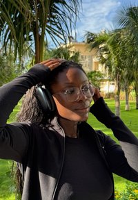 Kvinne med briller og svart langermet antrekk justerer svarte over-ear hodetelefoner utendørs med palmer og en gul bygning i bakgrunnen.