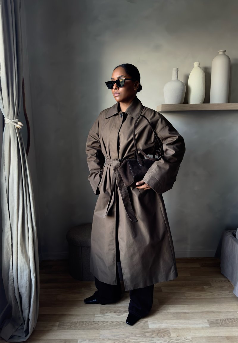 Femme en trench-coat marron oversize et lunettes de soleil, debout à l'intérieur, tenant un sac à main marron assorti, près d'un rideau et de vases décoratifs.