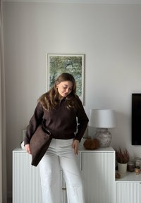 Brązowy sweter prążkowany, białe spodnie o szerokich nogawkach oraz brązowa torba o fakturze. Miękkie tekstury i neutralne kolory, z minimalistycznym tłem.