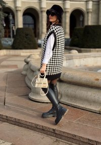 Czarnobiała kamizelka w wzór houndstooth w oversize'owym fasonie, biała koszula, czarne skórzane kozaki do ud oraz mała kremowa torebka. Czapka z czarnego futra i okulary przeciwsłoneczne.