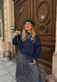 Marineblå sweatshirt med oversized ærmer, ternet nederdel med gennemsigtige lag, grøn beret og runde briller; holder et lille kamera og en kaffekop.