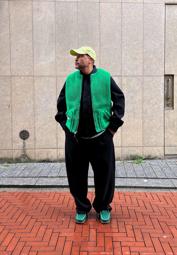 Colete de fleece verde por cima de uma camisa preta, combinado com calças largas pretas e sapatos verdes. Um boné verde limão completa o conjunto.