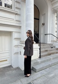 Bruine op maat gemaakte blazer over een zwarte fluwelen jurk, gecombineerd met pumps met spitse neus. Staande naast een gebouw met zuilen en trappen.