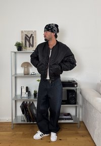 Zwarte bomberjack met een gladde textuur, gedragen over een wit T-shirt. Gecombineerd met losse donkere broeken en witte sneakers. Zwarte bandana.