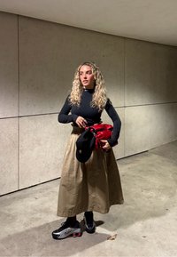 Svart langermet topp, beige midiskjørt og svarte joggesko. Holder en rød veske og har på seg en svart caps. Betongvegg i bakgrunnen.