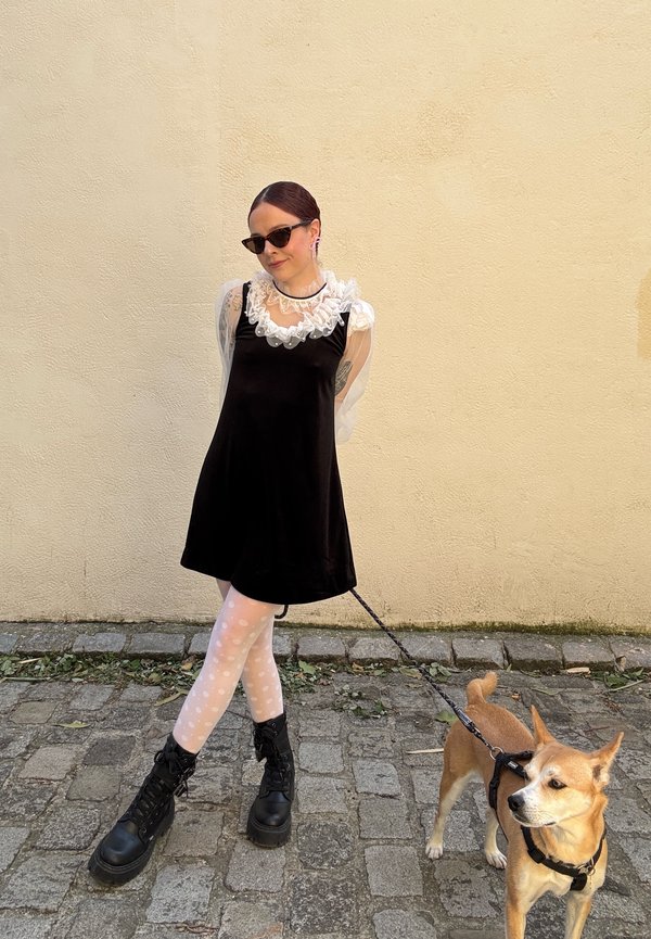 Vestido preto de veludo com colarinho de renda branca, meias de bolinhas e botas de combate pretas. Um cão castanho na trela está ao lado da pessoa.