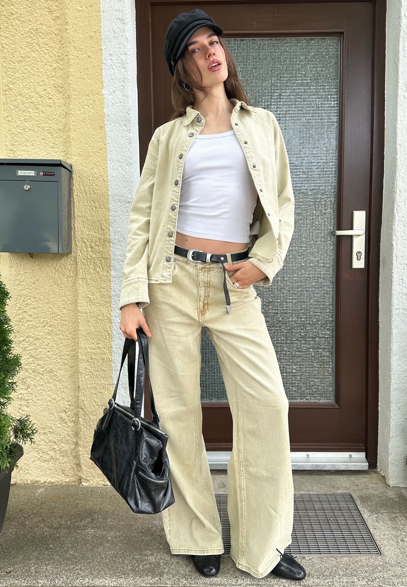Femme portant une veste et un pantalon beige, un crop top blanc, un chapeau noir, tenant un sac à main noir, debout devant une porte marron.
