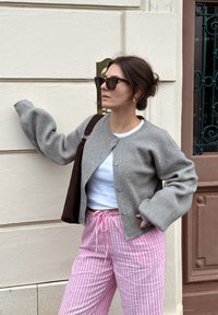 Sivi rebrasti kardigan s oversize rukavima, nosi se preko bijele bluze. Uparen s hlačama s ručnim vezanjem u ružičastim prugama i smeškom torbom. Tamne sunčane naočale naglašavaju.