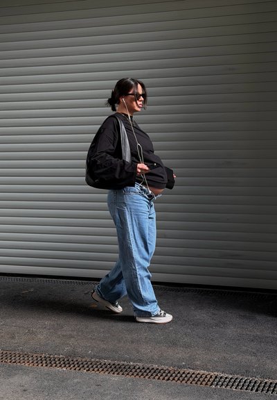 Mujer embarazada con gafas de sol, top negro y vaqueros camina junto a una persiana metálica gris, sosteniendo un teléfono con auriculares conectados.