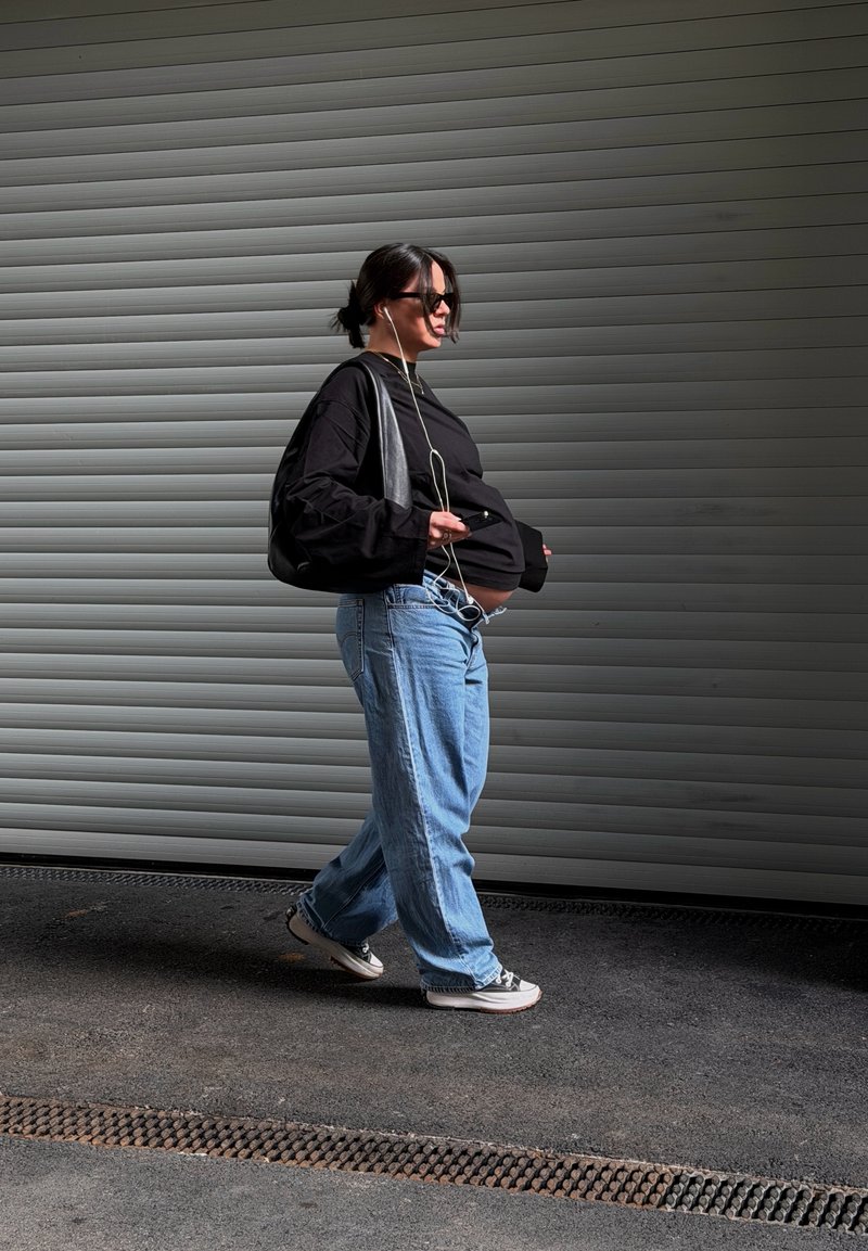 Raskaana oleva nainen, jolla on aurinkolasit, musta toppi ja farkut, kävelee harmaan metalliverhon ohitse ja pitää kädessään puhelinta, johon on kytketty kuulokkeet.