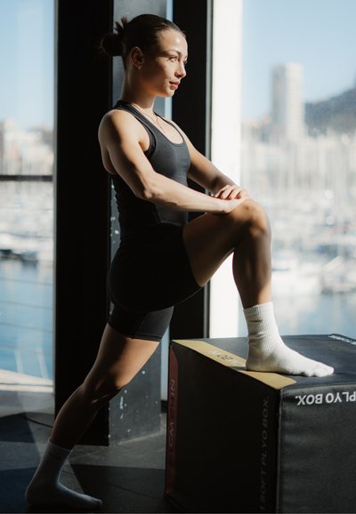 Mujer con ropa deportiva negra estirando una pierna sobre una caja de pliometría junto a una ventana grande con una marina visible afuera.