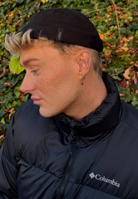 Svart puffjacka med hög krage, med en dragkedja och Columbia-logotyp. Svart keps på en ljushårig person, med ett guldörhänge i örat.