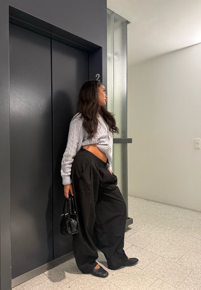 Mujer con un suéter de punto trenzado gris claro, pantalones negros de pierna ancha y zapatos planos negros, apoyada contra una pared cerca de un ascensor.