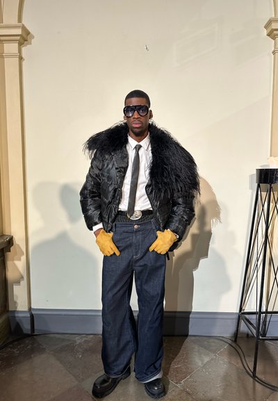 Hombre con chaqueta de piel negra, camisa blanca, corbata negra, gafas de sol grandes, guantes amarillos, vaqueros anchos y zapatos negros, de pie en el interior.