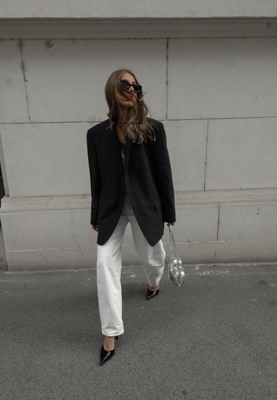 Blazer negro de gran tamaño, pantalones anchos blancos, tacones negros de punta y bolso plateado. Cabello largo, lleva gafas de sol oscuras. Fondo urbano.