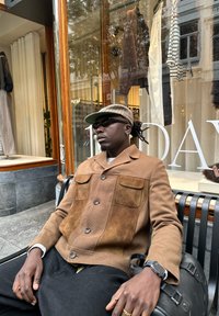 Giacca marrone con bottoni, davanti in suede, due tasche e maniche lunghe; abbinata a pantaloni neri. Seduto su una panchina vicino a una vetrina.