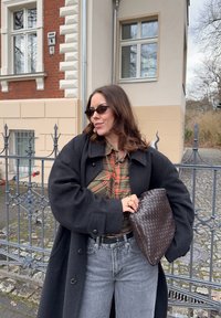 Juodas vilnonis paltas, languotas mygtukų marškinėliai (žali ir oranžiniai), aukšto liemens džinsai ir pintas rudas delninukas. Lauko miesto aplinka.