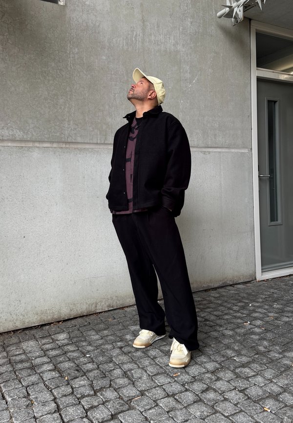 Un homme portant une veste et un pantalon noirs, une casquette beige et des baskets, debout sur des pavés, regardant vers le haut près d'un mur en béton gris et d'une porte.