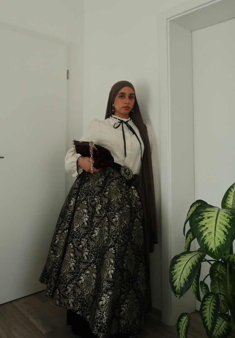 Longue jupe noire avec motifs floraux dorés, blouse blanche avec nœud ruban, et hijab marron. Tient une pochette avec une chaîne. Plante verte à proximité.