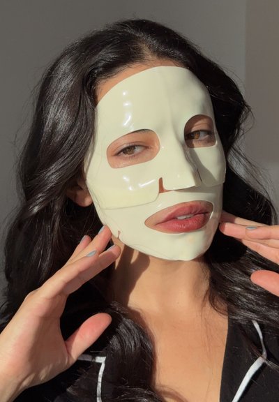 Mascarilla de hoja en beige claro con recortes para ojos y boca. El cabello oscuro del modelo cae alrededor de los hombros, las manos tocan la mascarilla con suavidad.