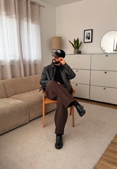 Hombre con gorra negra, gafas, chaqueta negra y pantalones marrones sentado con las piernas cruzadas en una silla de madera en una sala de estar beige con sofá y mueble.