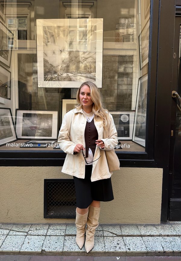 Mujer con el pelo largo y rubio, que lleva un abrigo beige y botas, está frente a una ventana de galería que exhibe fotografías en blanco y negro enmarcadas.