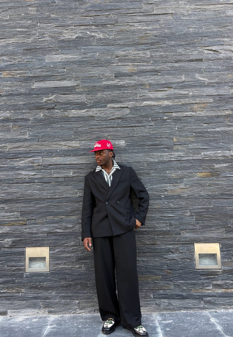 Homme en costume noir, chemise rayée, casquette rouge et chaussures à motifs, debout contre un mur en pierre grise texturé avec deux lumières carrées près du sol.