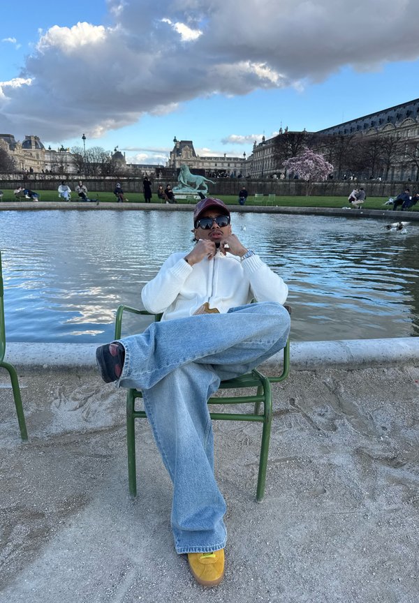 Mand iført solbriller og kasket, siddende med benene over kors på en grøn stol ved en stor rund dam i en park.