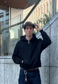 Schwarzer Kapuzenpullover, dunkle Jeans und herabhängende Schlüssel. Hält schwarze Kopfhörer auf dem Kopf, steht gegen eine Steinmauer mit Glaselementen.