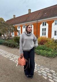 Světlý šedý pletený svetr s vysokým límcem, v kombinaci s černými džínami s širokými nohavicemi. Drží tkanou korálově růžovou kabelku s přívěskem ve tvaru bambulky.