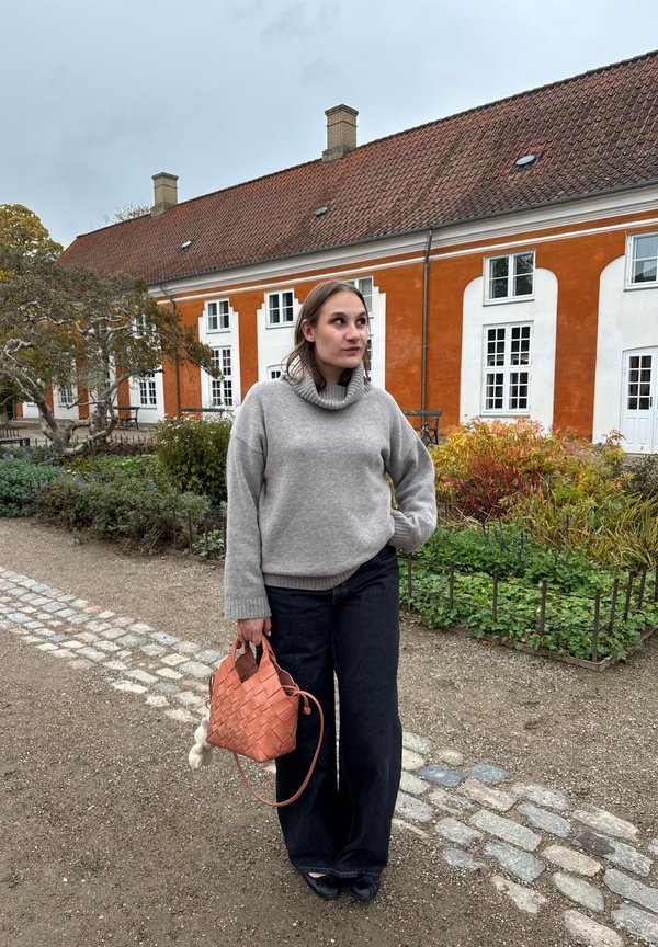 Světlý šedý pletený svetr s vysokým límcem, v kombinaci s černými džínami s širokými nohavicemi. Drží tkanou korálově růžovou kabelku s přívěskem ve tvaru bambulky.