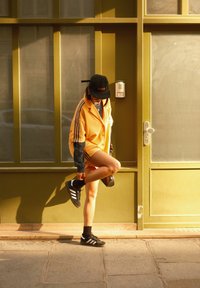 Nadrozmerná oranžová košeľa s modrými pruhmi cez čiernu dlhú rukávovú košeľu, v kombinácii s čiernymi šortkami a teniskami Adidas. Čierna šiltovka a detaily v podobe retiazok.