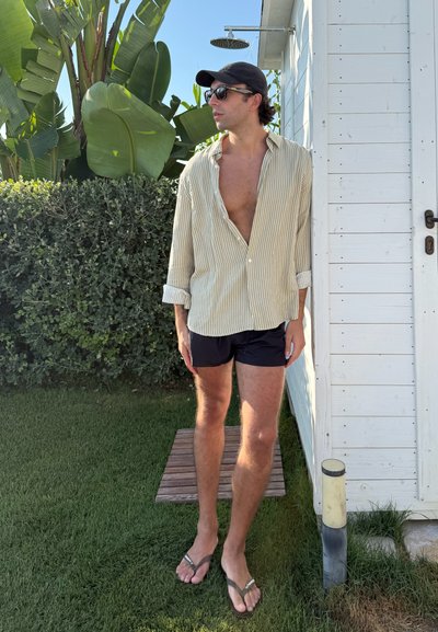 Camisa de botones a rayas claras, pantalones cortos azules y chanclas; de pie entre la vegetación cerca de un cobertizo blanco bajo un cielo azul.