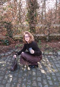 Femme portant des lunettes accroupie sur des pavés, tenant une tasse de café et un sac à main marron, à côté d'une clôture en bois et de feuillage sec.