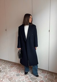 Γυναίκα με σκούρα μαλλιά μέχρι τους ώμους, φορώντας μακρύ μαύρο παλτό, λευκό πουλόβερ με ανάγλυφο πλέξιμο, μπλε τζιν, μαύρα παπούτσια και κρατώντας μια μαύρη τσάντα χεριού.