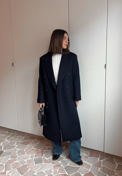 Mujer con cabello oscuro hasta los hombros, con un abrigo negro largo, suéter blanco de punto trenzado, jeans azules, zapatos negros y sosteniendo un bolso negro.